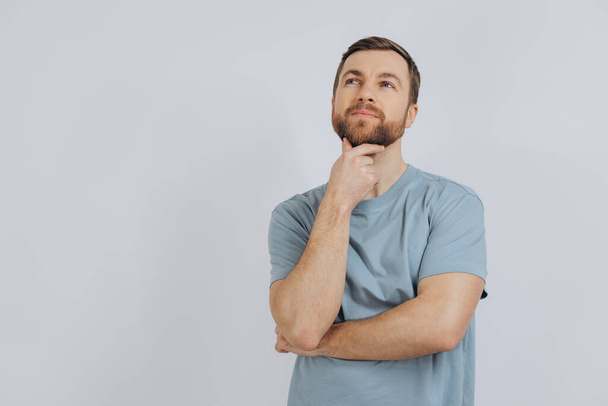 Retrato de un hombre barbudo moderno de mediana edad con una camiseta azul mostrando emoción pensando algo sobre un fondo blanco - Foto, Imagen