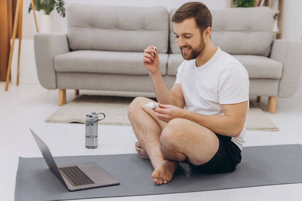 Atraktivní vousatý muž držící bezdrátová sluchátka poblíž notebooku a začínající ranní cvičení nebo jógu. - Fotografie, Obrázek