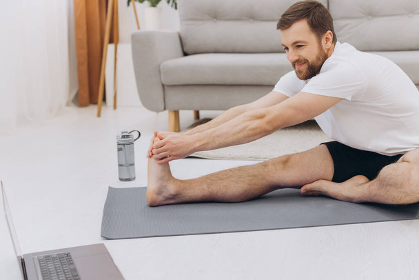 Handsome bearded man doing hamstring stretch exercise at home. - Zdjęcie, obraz