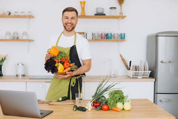 Portrait de l'homme heureux à la maison, homme cuisine salade de légumes en regardant la caméra et souriant, trancher des légumes, en utilisant un ordinateur portable pour la formation de cuisine en ligne - Photo, image