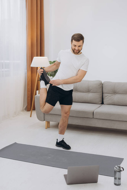 Positive athletic bearded middle-aged man stretching at home, using laptop, watching sport videos on Internet, having fitness class online, copy space. Healthy lifestyle, sport on self-isolation - Fotoğraf, Görsel