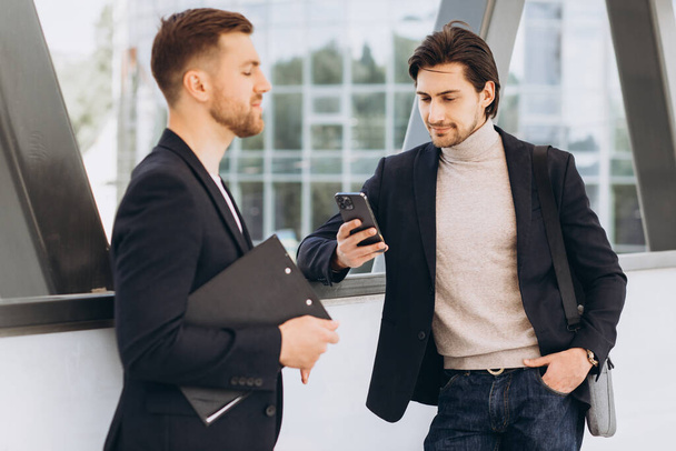 Two modern happy businessmen with phone and folder of documents discussing something against the background of urban offices and buildings - Foto, afbeelding