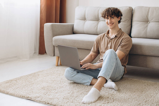 Portrait d'attrayant jeune homme aux cheveux bouclés utilisant un ordinateur portable tout en étant assis sur le canapé à la maison - Photo, image