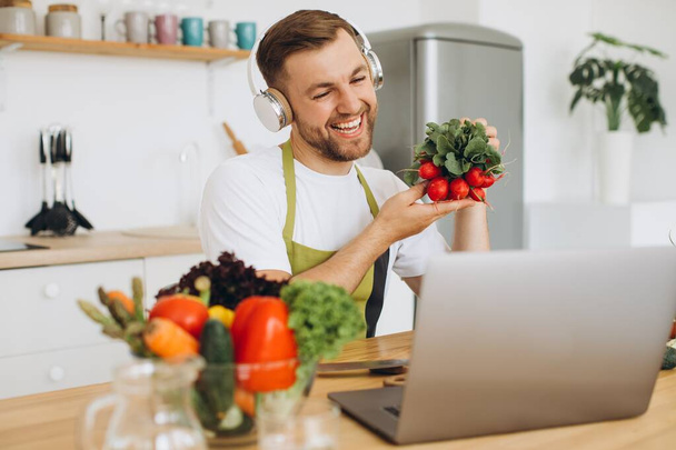 Šťastný muž ve sluchátkách sedí u kuchyňského stolu a připravuje salát, drží ředkvičku a ukazuje ji notebooku. - Fotografie, Obrázek