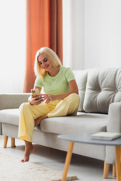 Attractive blonde middle-aged woman uses phone while sitting on sofa in living room at home - Valokuva, kuva