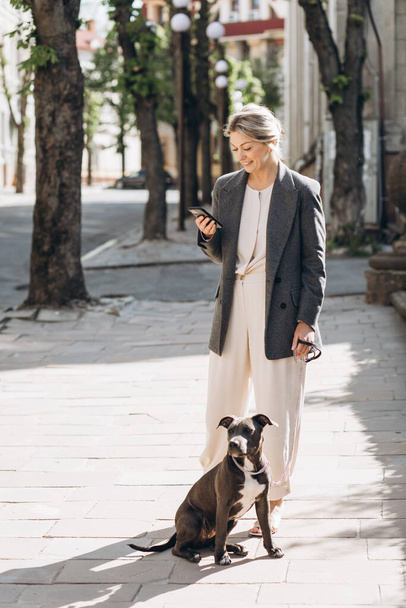 Gri ceketli olgun bir iş kadını köpeğini gezdirirken gülümsüyor ve telefonda konuşuyor. - Fotoğraf, Görsel