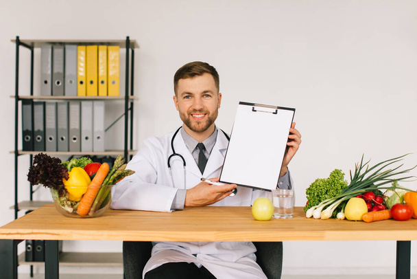 Szczęśliwy lekarz dietetyk siedzi w miejscu pracy przy biurku w biurze wśród świeżych warzyw i pokazuje folder z miejsca do kopiowania, koncepcja planu diety - Zdjęcie, obraz