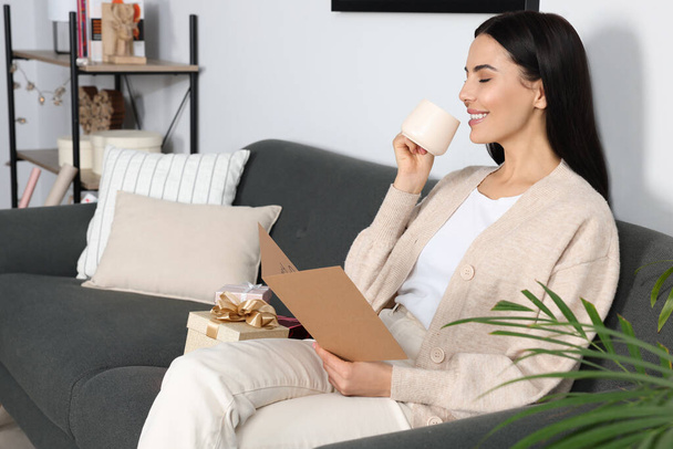 Happy woman reading greeting card while drinking coffee on sofa in living room - Photo, image