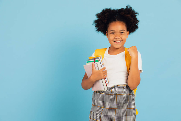 幸せなアフリカ系アメリカ人の女子高生が青い背景を指差して学校のコンセプトに戻る. - 写真・画像