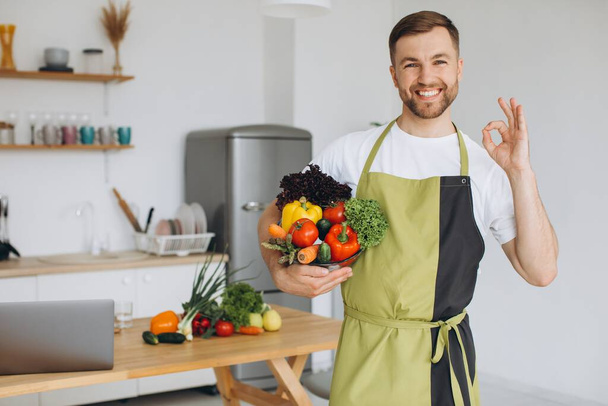 Πορτρέτο ενός ευτυχισμένου άνδρα κρατώντας ένα πιάτο φρέσκα λαχανικά στο φόντο της κουζίνας στο σπίτι - Φωτογραφία, εικόνα