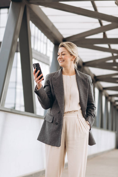 Portrait d'une belle femme d'affaires mature en costume et veste grise souriante et parlant au téléphone sur fond de bâtiments urbains et de bureaux modernes - Photo, image