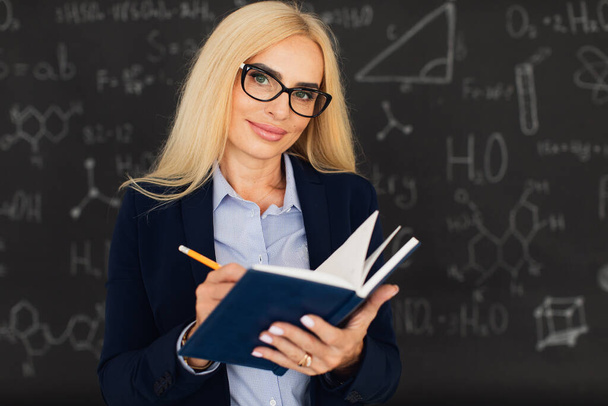 Porträt einer glücklichen blonden Lehrerin, die neben der Tafel im Klassenzimmer steht. - Foto, Bild