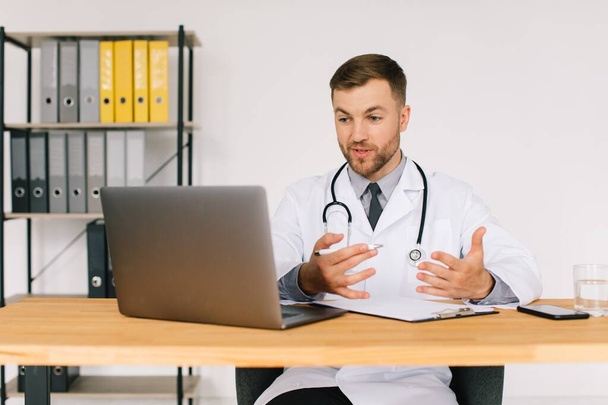 Heureux médecin masculin regardant un webinaire médical en ligne ou un séminaire de formation assis avec un ordinateur portable sur le lieu de travail. - Photo, image