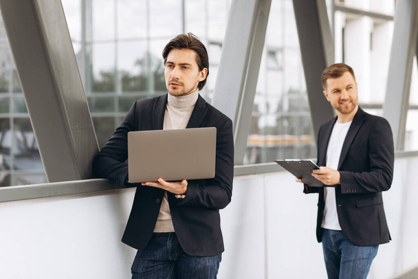 Due uomini d'affari felici moderni con laptop e documenti che discutono di qualcosa sullo sfondo di uffici e edifici urbani - Foto, immagini
