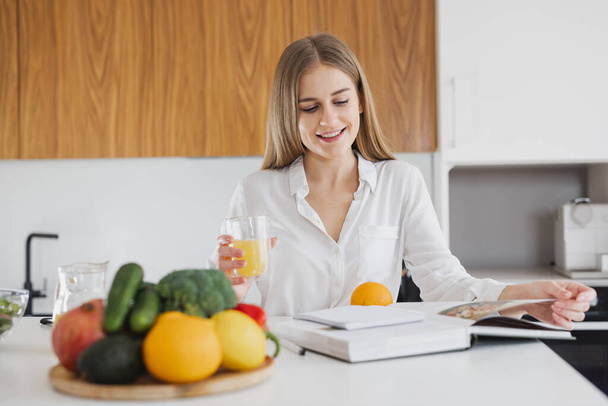 Cute blonde woman is looking at a recipe book and drinking juice in the kitchen - Foto, immagini