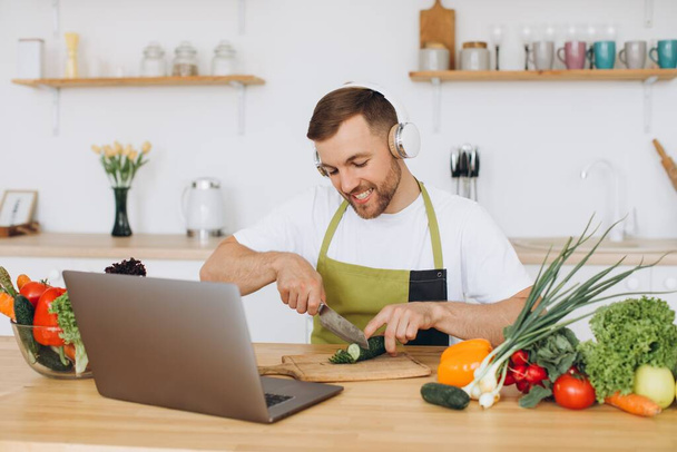 Homem feliz em fones de ouvido preparando salada na cozinha de legumes frescos e olhando para laptop. - Foto, Imagem