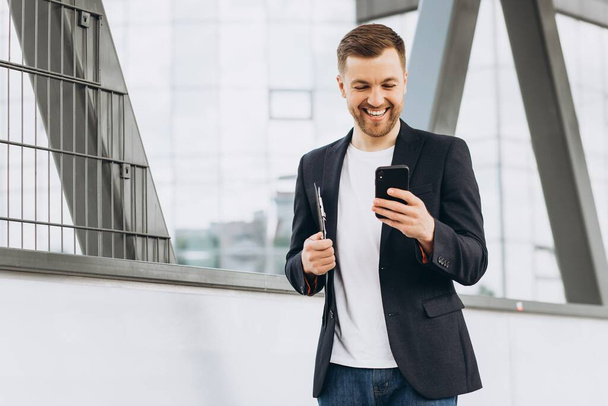 Ritratto di felice uomo d'affari moderno in giacca e cravatta che cammina sorridendo e leggendo o scrivendo messaggi al telefono sullo sfondo di edifici urbani e uffici. - Foto, immagini