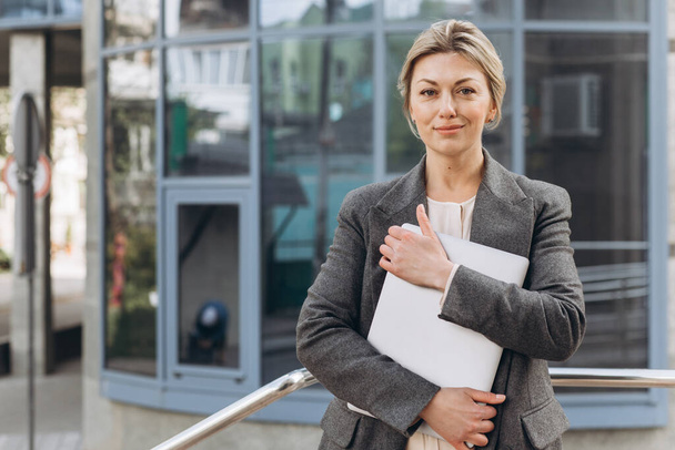 Portrait of a mature business woman smiling with emotions and holding a laptop on the modern urban and office buildings background - Foto, Bild