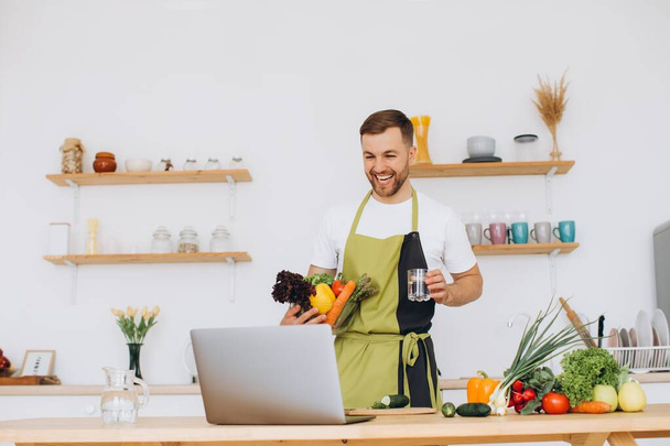 Πορτρέτο του ευτυχισμένος άνθρωπος στο σπίτι, άνθρωπος μαγείρεμα σαλάτα λαχανικών κοιτάζοντας κάμερα και χαμογελώντας, τεμαχίζοντας λαχανικά, χρησιμοποιώντας φορητό υπολογιστή για online προπόνηση μαγειρικής - Φωτογραφία, εικόνα