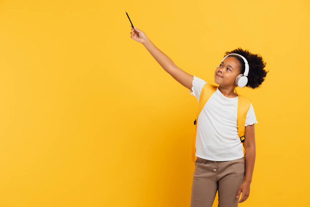Gelukkig Afrikaans amerikaans schoolmeisje in hoofdtelefoon met rugzak tonen iets met de hand in de klas op gele achtergrond, kopieer ruimte. - Foto, afbeelding