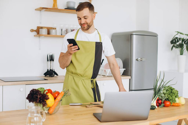 Atractivo joven está cocinando en la cocina con un ordenador portátil en la mesa, hablando en un teléfono inteligente. - Foto, imagen