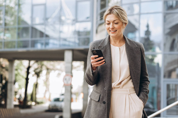 Portrait d'une belle femme d'affaires mature en costume et veste grise souriante et parlant sur un téléphone sur le fond des bâtiments urbains et de bureaux modernes - Photo, image