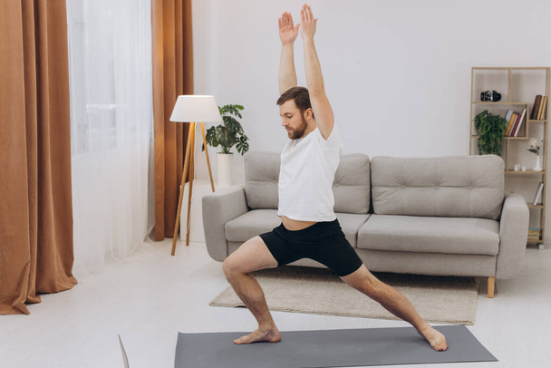 Positive athletic bearded middle-aged man stretching at home, using laptop, watching sport videos on Internet, having fitness class online, copy space. Healthy lifestyle, sport on self-isolation - Foto, afbeelding