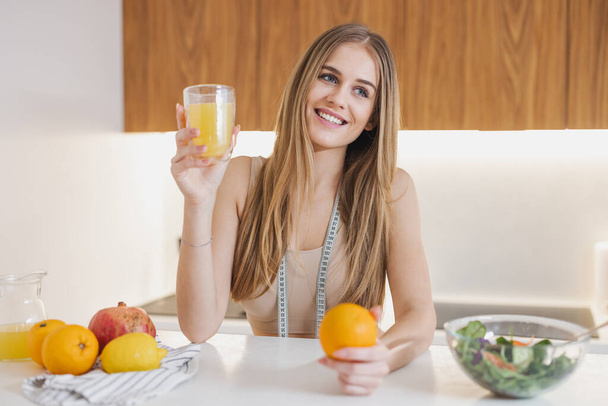 Ευτυχισμένη ελκυστική γυναίκα χιλιετηρίδας σε αθλητικά ρούχα με ταινία μέτρησης κρατώντας φρέσκο χυμό και πορτοκάλι για την ανάκτηση ενέργειας μετά την προπόνηση στην κουζίνα - Φωτογραφία, εικόνα