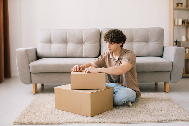 Happy young smiling curly man opening box with ordered goods gifts, presents at home on couch. Online shopper male customer opening online shop parcel. - Photo, Image