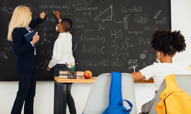 Female teacher teaches children at school near the blackboard. Quarantine, mask mode, covid-19. Back to school. - Φωτογραφία, εικόνα