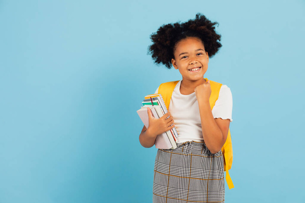 Happy Afro-Amerikaanse schoolmeisje wijzen cool op blauwe achtergrond, terug naar school concept. - Foto, afbeelding