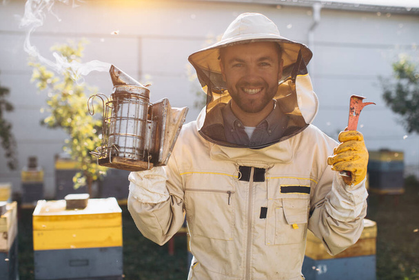 Portret van een gelukkige mannelijke imker die werkt in een bijenstal bij bijenkorven met bijen. Verzamel honing. Bijenhouder op de bijenstal. Bijenteeltconcept. - Foto, afbeelding