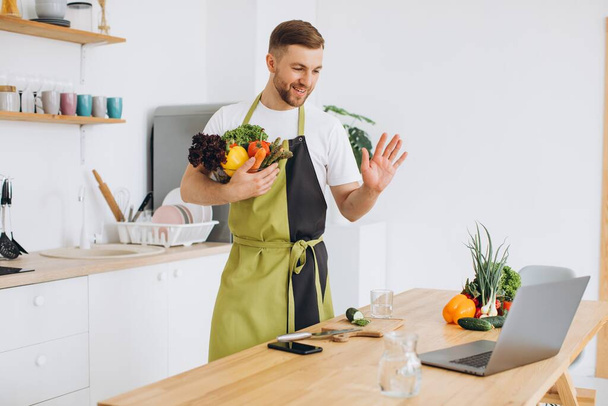 Portrait of happy man at home, cooking vegetable salad looking at camera and smiling, makes a greeting gesture with hand, using laptop for online cooking training - Photo, Image