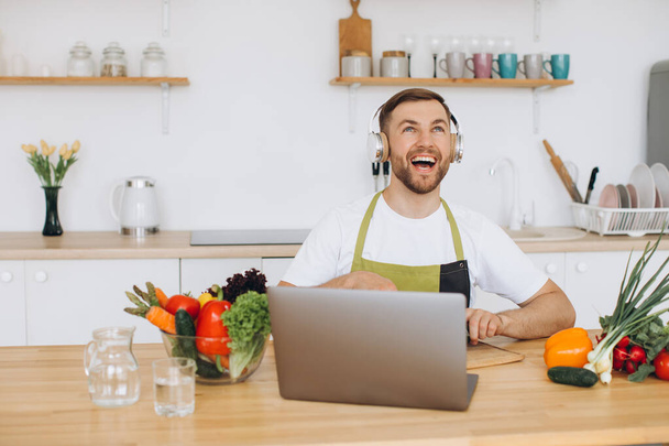 Happy man in headphones preparing salad in kitchen from fresh vegetables and looking at laptop. - 写真・画像