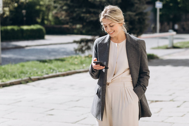 Madura rubia mujer de negocios en una chaqueta gris sonriendo caminando por la ciudad y sosteniendo un teléfono - Foto, imagen