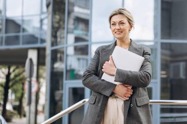 Portrait of a mature business woman smiling with emotions and holding a laptop on the modern urban and office buildings background - Zdjęcie, obraz