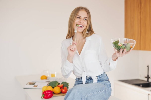 Щаслива і усміхнена блондинка сидить на кухні і насолоджується домашнім салатом серед інгредієнтів приготування їжі
. - Фото, зображення