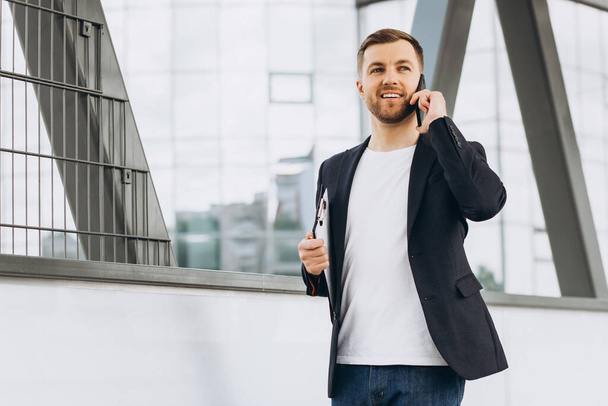 Portrait of happy modern male businessman in suit holding folder with documents and talking by phone against the background of urban buildings and offices. - 写真・画像