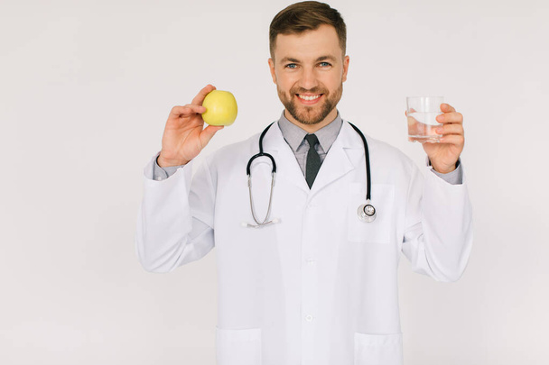 The happy male nutritionist doctor with stethoscope smiling and holding water and apple on white background, diet plan concept - Photo, Image