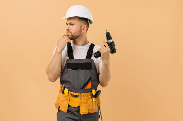 Positiver männlicher Reparateur zu Hause, der einen Schraubenzieher in der Hand hält und auf beigem Hintergrund denkt - Foto, Bild