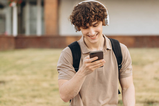 attrayant frisé jeune homme université ou étudiant avec téléphone marche autour du campus - Photo, image