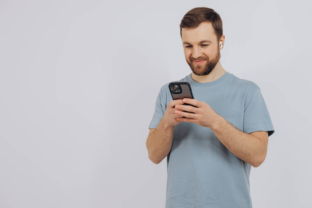 Portrait of a modern millennial man with a beard holding a smartphone and listening to something in wireless headphones on a white background with copy space - 写真・画像