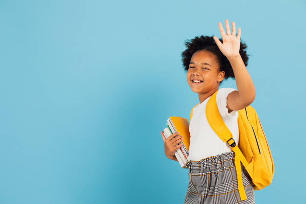 Happy African American schoolgirl makes a welcome hand gesture on blue background, back to school concept. - Foto, Imagen