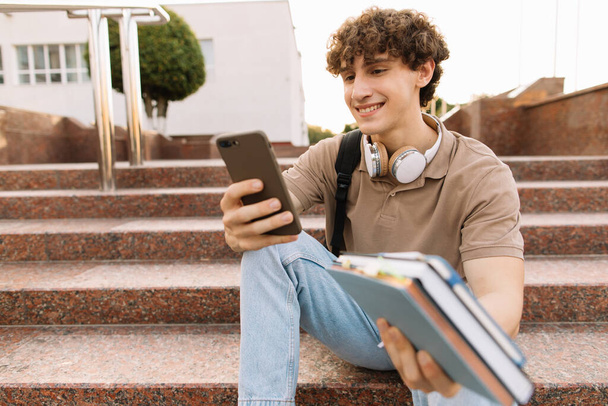 Çekici kıvırcık saçlı genç adam üniversite ya da üniversite öğrencisi merdivenlerde oturuyor ve akıllı telefon arıyor. - Fotoğraf, Görsel