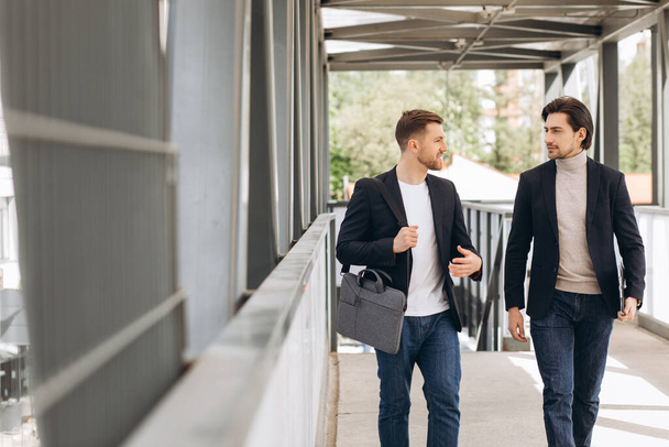 Два современных счастливых бизнесмена идут по городскому мосту и обсуждают что-то на фоне городских офисов и зданий - Фото, изображение