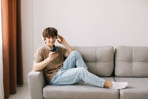 Portrait of young happy curly haired man using smartphone with headphones while sitting on sofa in living room and resting - Photo, image
