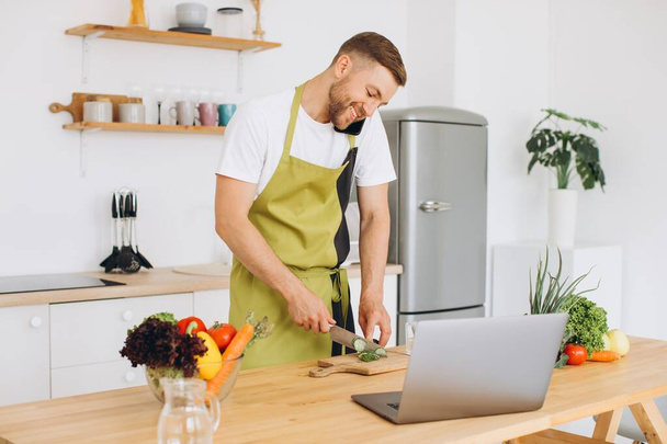 Jeune homme attrayant cuisine dans la cuisine avec un ordinateur portable sur la table, parler sur un smartphone. - Photo, image