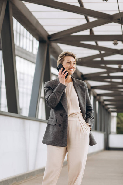 Πορτρέτο μιας όμορφης ώριμης γυναίκας με κοστούμι και γκρι σακάκι χαμογελώντας και μιλώντας στο τηλέφωνο σε ένα σύγχρονο αστικό και γραφείο κτίρια φόντο - Φωτογραφία, εικόνα