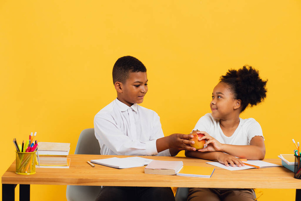 幸せなアフリカ系アメリカ人の女子高生と女子高生が机の上に座り、黄色い背景でリンゴを分かち合っています。学校のコンセプトに戻る. - 写真・画像