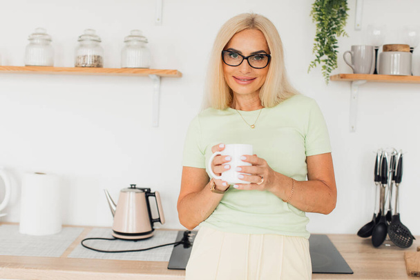 Σύγχρονη ελκυστική μεσήλικη γυναίκα πίνοντας καφέ στην κουζίνα στο σπίτι - Φωτογραφία, εικόνα